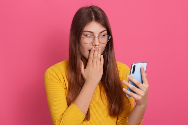 Picture of emotional shocked impressed young model holding smartphone, looking at device screen attentively, covering her mouth with hand, being surprised by news. People and emotions concept.