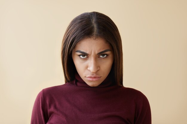 Picture of evil young dark skinned brunette woman having sinister appearance, staring from under her frowned eyebrows, grabbing lips furiously, her look full of anger and irritation