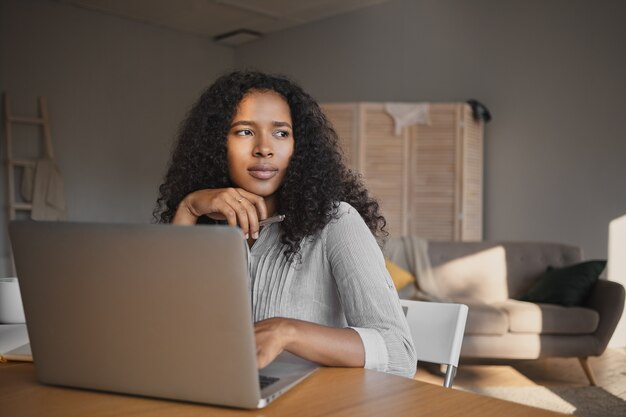 Picture of fashionable attractive young dark skinned woman freelancer in shirt sitting at workplace at home and using high speed wirless internet connection on laptop, having thoughtful look