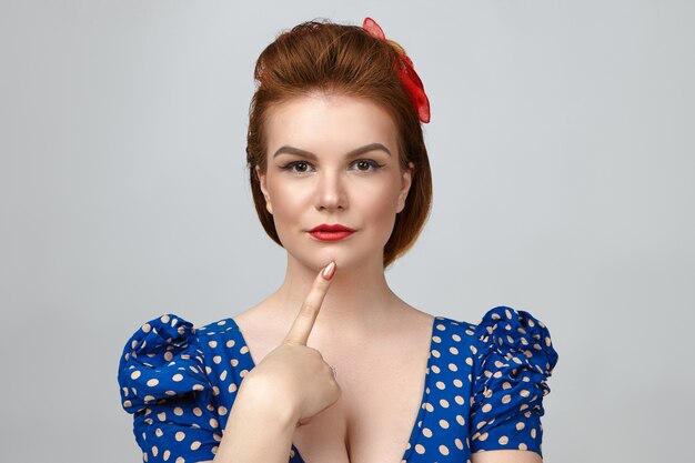 Picture of fashionable glamorous young red haired female dressed in pin up style posing isolated in studio, holding fore finger on her chin and staring at camera with pensive thoughtful look