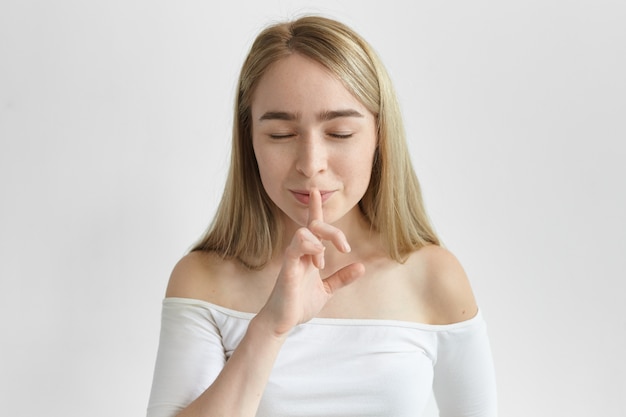 Picture of fashionable young woman with fair hair and freckles shushing, closing eyes and keeping index finger at her mouth, asking to keep her secret. Secrecy and confidential information