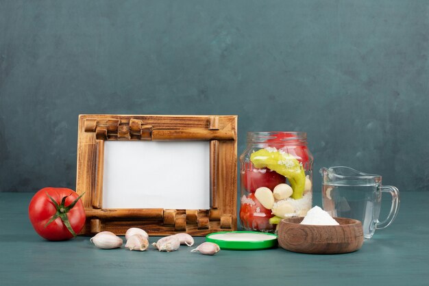 Picture frame, pickled in glass jar, salt and fresh vegetables on blue table. 