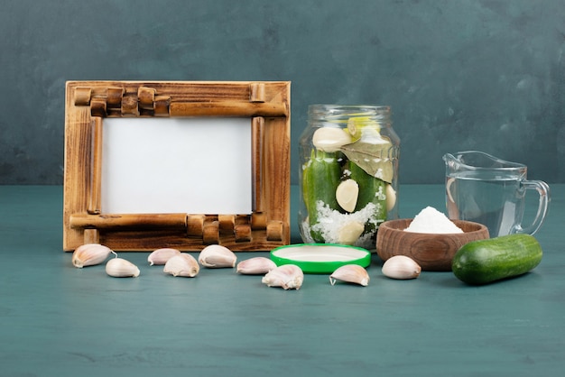 Picture frame, pickled vegetables in glass jar and salt bowl on blue surface with fresh cucumber and garlic. 