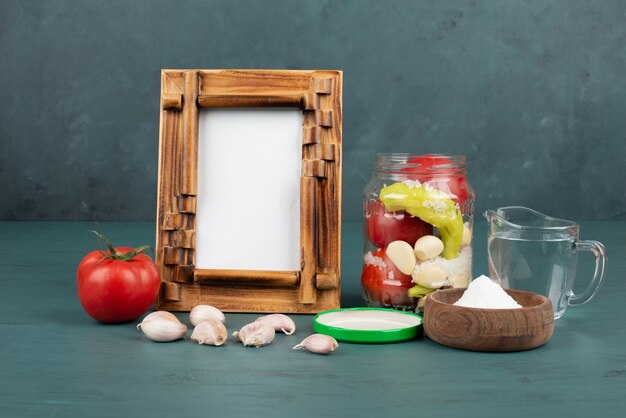 Picture frame, pickled vegetables in glass jar and salt bowl on blue surface with fresh tomato and garlic. 