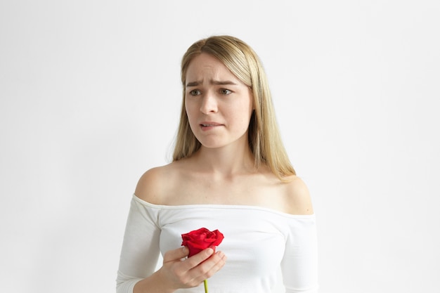 Foto di giovane e bella signora frustrata in posa con una rosa rossa del suo amante, preoccupata e sconvolta, mordendosi le labbra