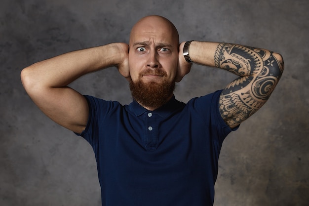 Free photo picture of frustrated emotional european man with shaved head and fuzzy beard stylish hair grimacing, going to cry because of loud noise or argument with his girlfriend, covering his ears with hands