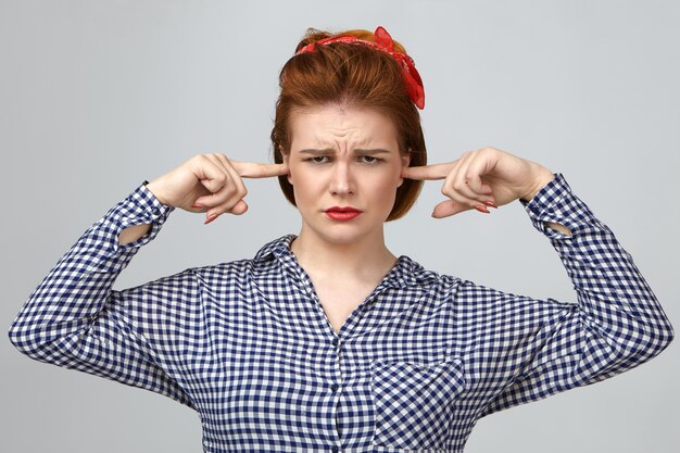 Picture of frustrated young housewife having painful fed up look, frowning and plugging ears with fingers, being stressed with noisy children