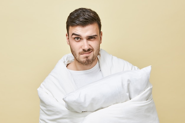 일찍 일어나야한다는 스트레스를 받고, 손에 베개와 화난 표정이있는 하얀 부드러운 담요로 싸여있는 좌절 된 젊은 형태가없는 남자의 사진. 침구 컨셉