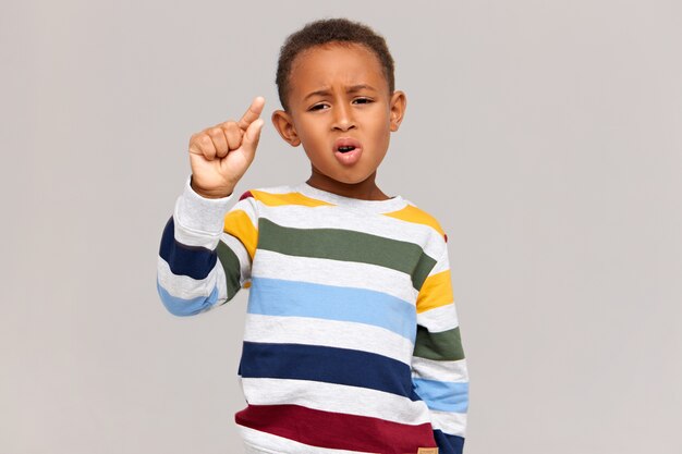 Picture of funny dark skinned schoolboy in stylish striped jumper having disappointed facial expression, making gesture with fingers as if holding something very tiny. Discount, sale and small price