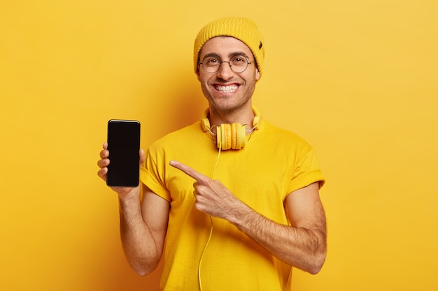 Picture of glad handsome man shows display of smartphone, points at modern gadget