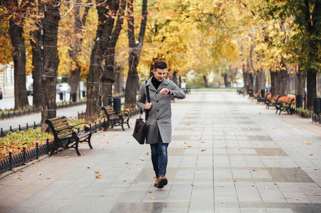 Picture of good-looking caucasian man in coat with bag strolling in city park, and looking at his watch