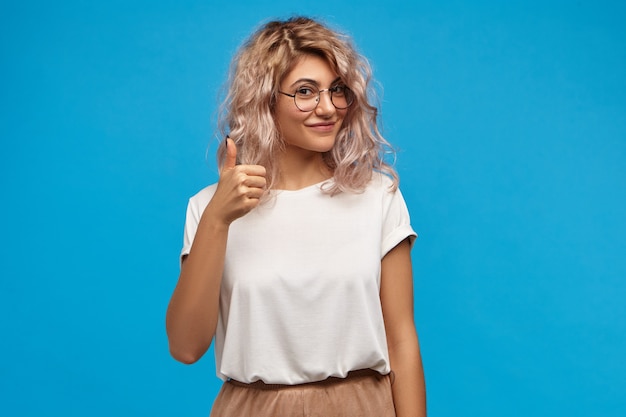 Foto di bella giovane femmina caucasica amichevole che indossa abiti eleganti e occhiali rotondi che fa il gesto di approvazione, mostrando il pollice in alto segno alla macchina fotografica e sorridendo felicemente