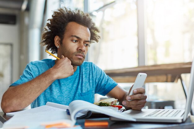 Picture of good-looking smart dark-skinned student in blue t-shirt working on course paper at college canteen, having sandwich and checking newsfeed via social networks on cell phone.