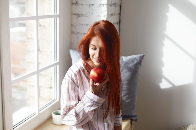 Фотография великолепной улыбающейся молодой рыжеволосой девушки в стильной пижаме, позирующей у большого окна в своей комнате, держащей большое спелое красное яблоко, возбужденной ожидаемым взглядом, наслаждающейся вкусными здоровыми фруктами