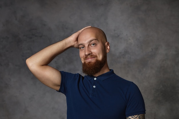 Picture of handsome attractive young bearded man wearing stylish polo shirt  with flirty expression, scratching his shaved bald head, feeling shy while talking to beautiful woman