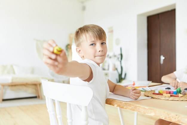 Picture of handsome Caucasian schoolboy with fair hair and blue eyes sitting at wooden table and making figures using modeling clay, showing yellow model. Selective focus on child's face