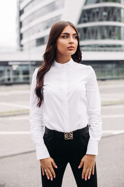 Foto gratuita immagine di una bella donna caucasica con lunghi capelli ondulati scuri in una camicia bianca, pantaloni neri e tacchi in posa per la fotocamera