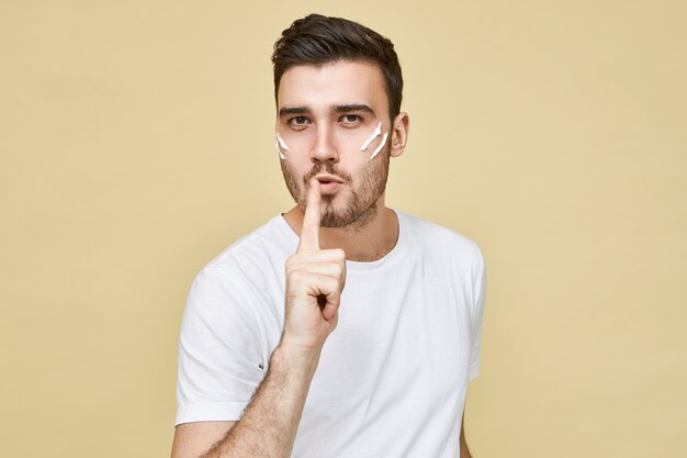 Picture of handsome confident young male in white t-shirt keeping index finger at his lips as if blowing at gunpoint after shooting making shush sign. Secrecy and silence gesture