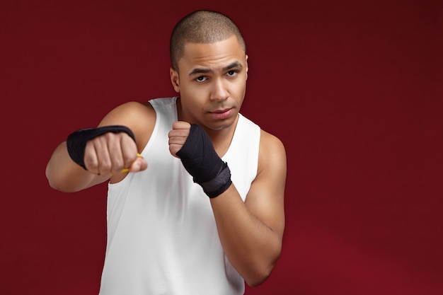 ボクシングの試合中に相手を殴る準備ができて、彼の前に拳を保ち、空白の壁でジムに立っている筋肉の腕を持つハンサムな強い運動の若い混血男ボクサーの写真