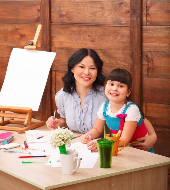 Изображение счастливой матери и дочери, улыбающихся в камеру, сидя за столом и рисуя картинки