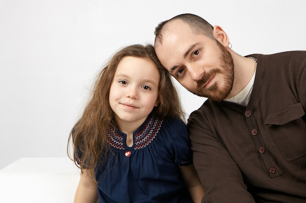 Picture of happy young single father with stylish beard smiling at camera with his charming female child, posing against white studio wall background with copyspace