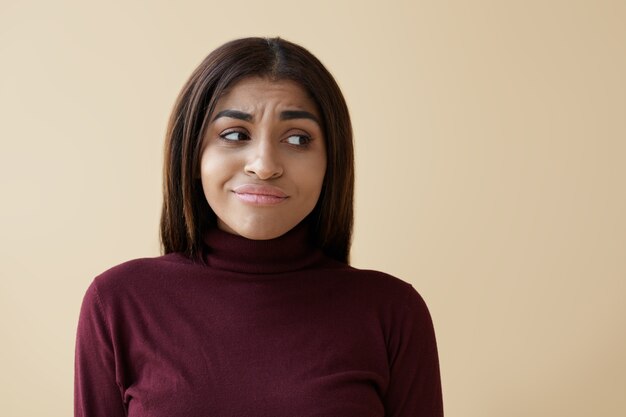 Picture of hesitant young mixed race woman with dark long hair frowning and looking sideways with perplexed doubtful facial expression