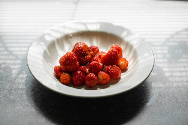 Picture of juicy fresh ripe red strawberries in a white ceramic plate on the table under bright sunlight in a village