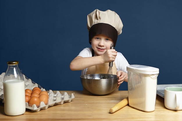 Изображение маленького мальчика в фартуке и кепке от шеф-повара, взбивающего ингредиенты в металлической миске во время приготовления блинов, печенья или другого теста, стоящего за кухонным столом с яйцами, молоком, мукой и скалкой