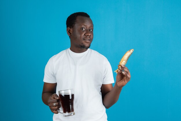 青い壁にワインのガラスのマグカップと干物を保持している白いTシャツの男の写真