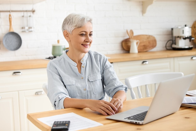Picture of modern beautiful retired female using wirless internet connection on portable computer, sitting at table in stylish kitchen interior, looking away with thoughtful pensive facial expression