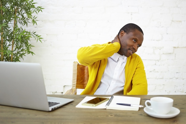 Picture of modern fashionable young dark skinned businessman rubbing neck, feeling frustrated and uncertain about something, sitting at workplace with open laptop, diary, mug and mobile phone on desk