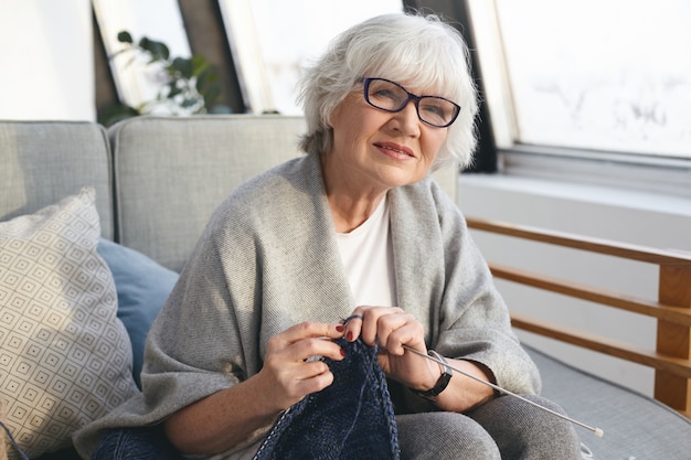 Picture of neat retired woman wearing wide scarf and eyeglasses knitting warm jumper for her daughter. Attractive senior female knitter working from home, handmaking winter clothes for sale