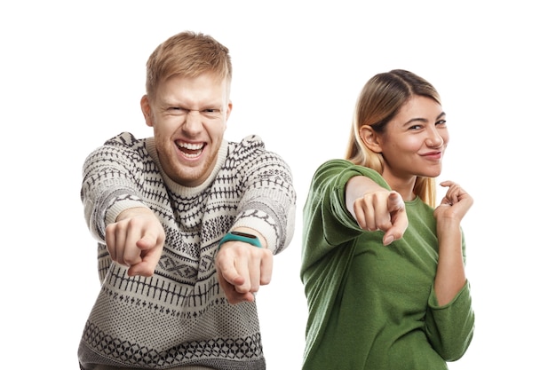 Picture of overjoyed ecstatic young Caucasian couple bearded man and blonde woman standing next to each other, having excited happy looks and pointing fingers, choosing you