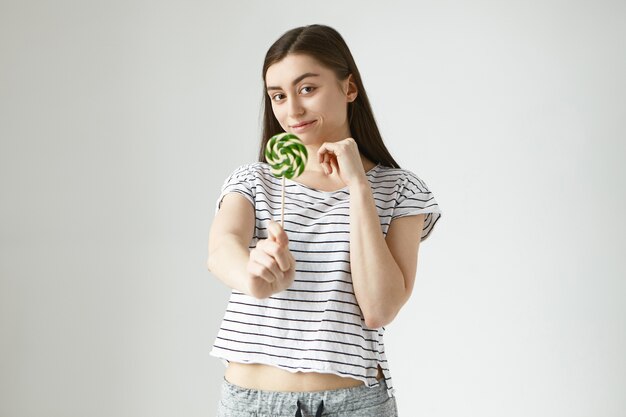 Picture of positive happy 20 year old woman with shiny dark hair reaching out hand with spiral colorful sweet hard candy, offering you to have it. People, food, nutrition, diet and sweets concept