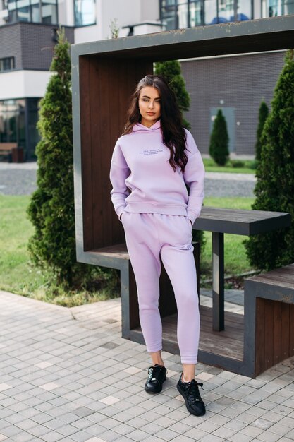 Picture of pretty caucasian female in purple sport suit and black trainers keeps her hands in the pockets