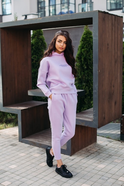 Picture of pretty caucasian female in purple sport suit and black trainers keeps her hands in the pockets