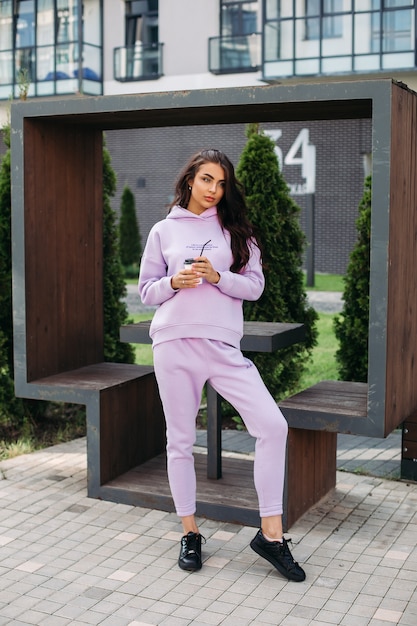 Picture of pretty caucasian female in purple sport suit and black trainers keeps her hands in the pockets