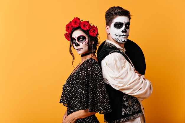 Picture of proud Mexican couple in traditional clothes with painted faces. Girl with roses in her hair poses with young man with sombrero behind his back.