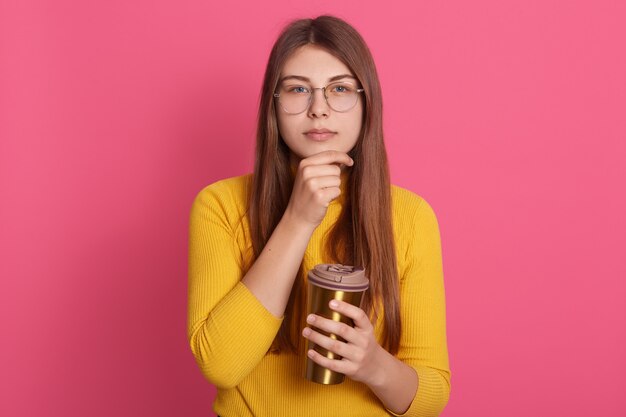 Picture of serious thoughtful young model holding mug with drink, looking directly  having pensive facial expression