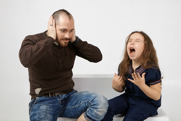 Free photo picture of shocked young unshaven male in stylish clothes covering ears because of naughty spoiled female child who is crying and shouting loudly.