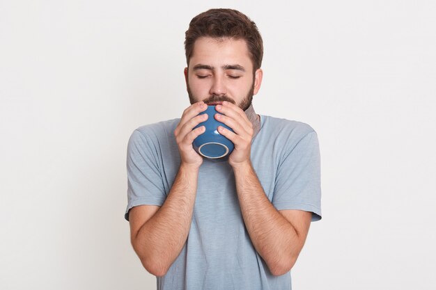 Picture of sleepy dreamy peaceful young man closing eyes, drinking coffee, posing isolated over white wall in 