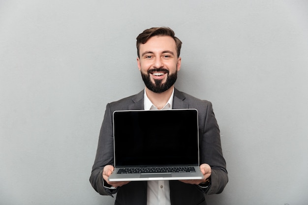 Picture of smiling bearded man holding silver personal computer showing black screen and looking on camera, isolated over gray wall