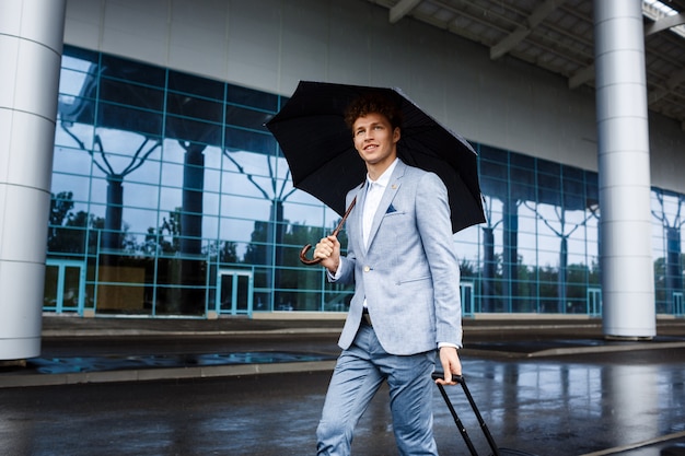 Picture of smiling young redhaired businessman holding umbrella and suitcase in rain at airport