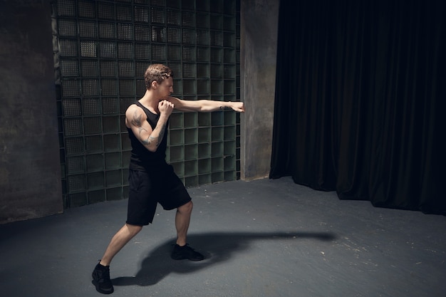 Picture of stylish fit Caucasian guy with muscular tattooed shoulders boxing in empty room reaching out one hand, mastering punches while preparing for fight. People, healthy lifestyle and sports