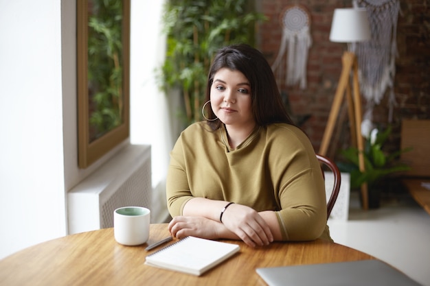 Picture of stylish overweight young female designer sitting at coworking place and drinking coffee, thinking over concept of new project, sketching in diary, having pensive thoughtful look