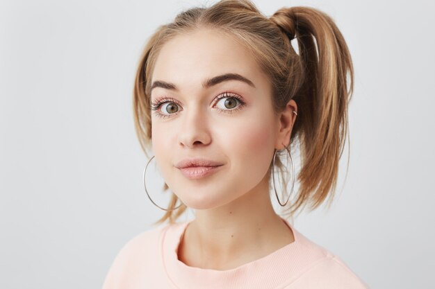 Picture of stylish trendy-looking teenage girl of European appearance wearing pastel pink loose sweater, with two ponytails and round earings, looking with appeal . Face expression