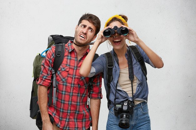 Picture of tired bearded man tourist carrying heavy backpack and cheerful excited woman with photo camera looking for place for camping using binocular during hiking trip together. People and adventure