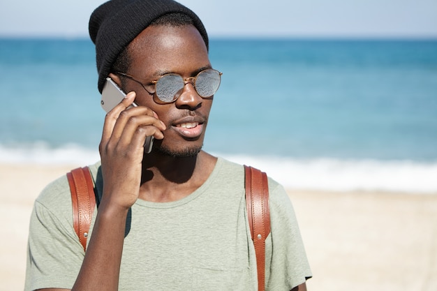 Картина модно выглядящий черный мужчина турист с рюкзаком, в шляпе и в темных очках в солнечную погоду, разговаривает по мобильному телефону