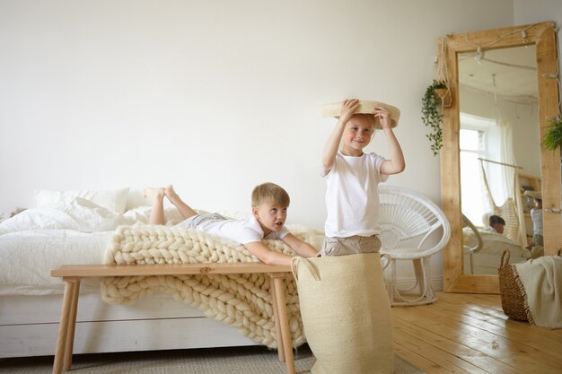 Picture of two adorable Caucasian schoolboys having fun indoors, playing active games together in parents' bedroom, feeling happy and carefree. Cute male children entertaining themselves at home