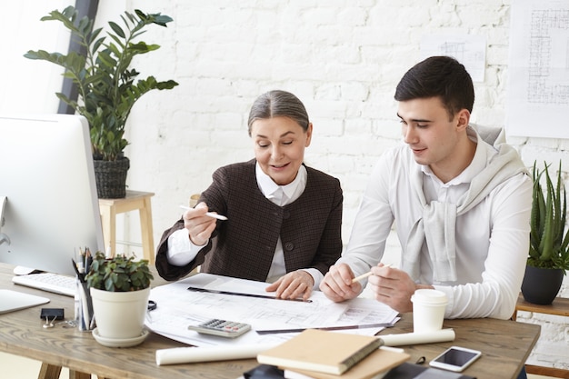 Picture of two cheerful professional designers mature woman and young man working on residential building project together, sitting at desk, discussing functional space creation and decoration ideas